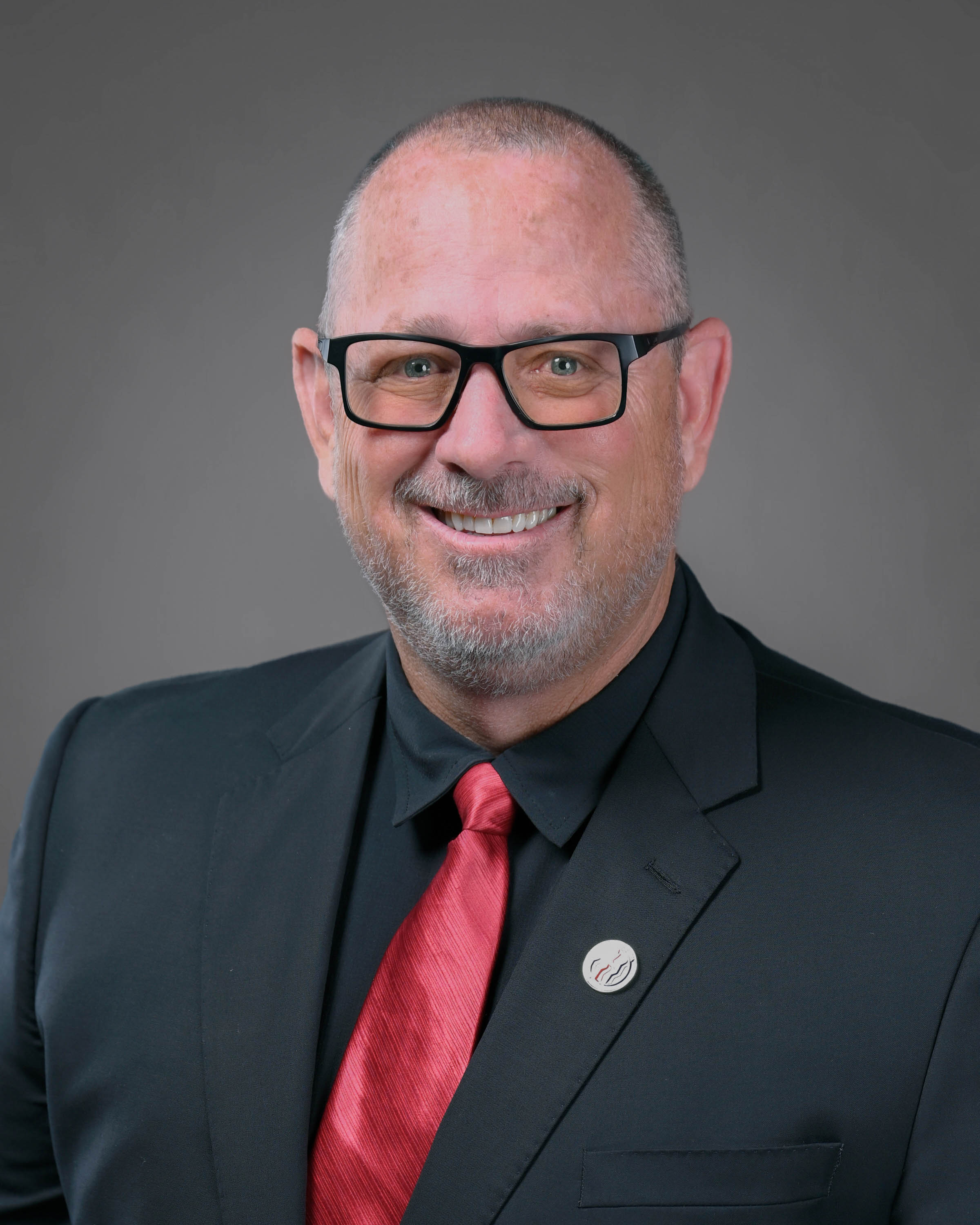 Man with short hair and beard wearing eyeglasses and a dark suit with a red tie